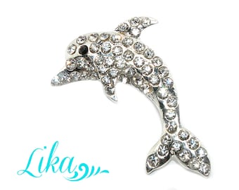 Puede incluir: Broche de delfín plateado incrustado con pedrería transparente. El delfín está en una pose de natación, con su cola y aleta dorsal visibles. El broche está sobre un fondo blanco.