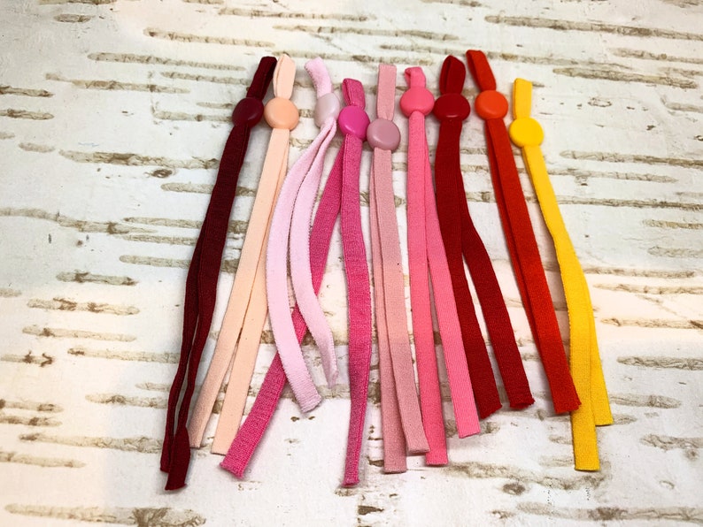 K&ouml;nnte beinhalten: Sortiment an bunten elastischen Maskenverl&auml;ngerungen mit Plastikkn&ouml;pfen. Die Verl&auml;ngerungen sind in Rot-, Rosa-, Orange- und Gelbt&ouml;nen.