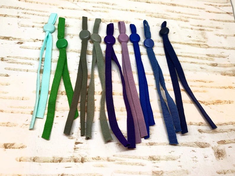 K&ouml;nnte beinhalten: Sortiment von 10 bunten elastischen Maskenverl&auml;ngerungen mit verstellbaren Schiebern. Die Verl&auml;ngerungen sind aus Stoff und in verschiedenen Farben erh&auml;ltlich, darunter Gr&uuml;n, Blau, Lila und Grau.
