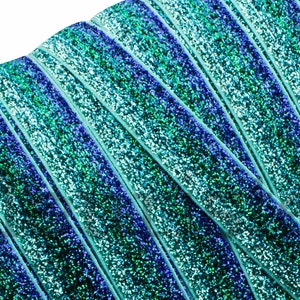 May include: Close-up of turquoise, blue, and green glitter ribbon. The ribbon is folded and layered, creating a textured, iridescent effect.