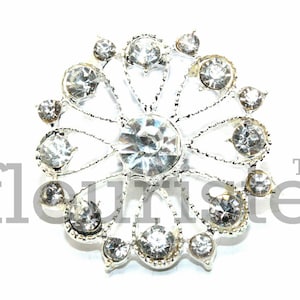 May include: Silver-toned brooch with a circular design featuring a central rhinestone surrounded by smaller rhinestones in a floral pattern.