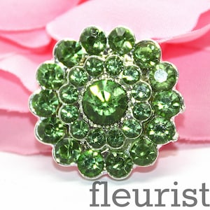 May include: A silver-toned flower-shaped embellishment with green rhinestones. The rhinestones are arranged in a circular pattern, creating a floral design.