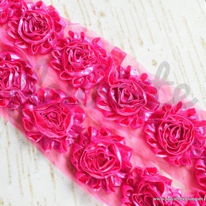 May include: Bright pink floral ribbon featuring dimensional fabric roses. The roses are arranged in a row on a sheer pink mesh backing. The ribbon is suitable for embellishments, crafts, and decorative projects.