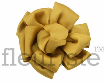 May include: Mustard yellow fabric bow with multiple layers and folds. The bow is centered on a white background. The word "fleuriste" is printed in light gray on the lower left side of the bow.