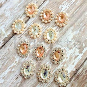 May include: An assortment of decorative embellishments with oval-shaped, faceted gemstones in peach and clear colors. Each gem is surrounded by a ring of small pearls or clear rhinestones, set on a light wood surface.