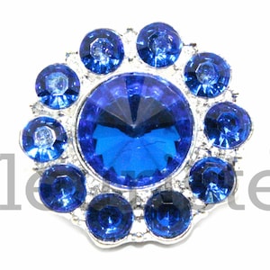 May include: A silver-toned, flower-shaped embellishment with a large, blue rhinestone in the center and smaller blue rhinestones surrounding it.