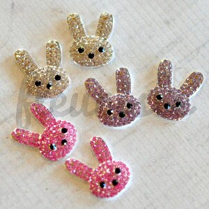 May include: Six sparkly rabbit-shaped embellishments. Three are pink with black eyes, two are light brown with black eyes, and one is purple with black eyes.