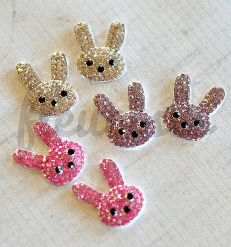 May include: Six sparkly rabbit-shaped embellishments. Three are pink with black eyes, two are light brown with black eyes, and one is purple with black eyes.
