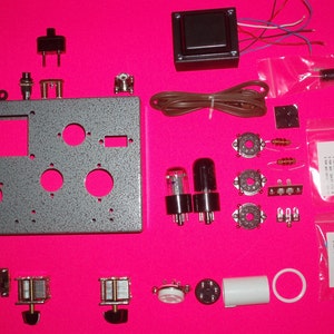 May include: A kit for building a radio, containing a variety of components including a chassis, valves, resistors, capacitors, and a transformer. The kit is laid out on a pink surface.