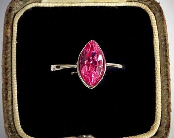 May include: A marquise-cut pink gemstone ring set in a silver band, displayed in an open vintage jewelry box with a black interior. The ring's gemstone is a vibrant pink color, and the box has a worn, leather-like exterior.
