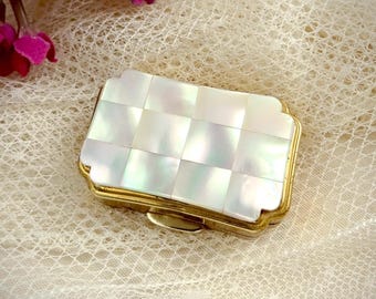 May include: A rectangular pill box with a gold-toned metal frame and a lid featuring a grid of iridescent white mother-of-pearl tiles. The box is positioned near a cluster of small, vibrant pink flowers with green stems.