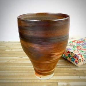 May include: A handcrafted ceramic cup with a wide top and a tapered base. The cup displays bands of brown and orange tones. A floral patterned box sits on a wooden surface beside the cup.