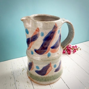 May include: A speckled ceramic jug with a handle and spout. The jug is decorated with blue dots and abstract shapes in shades of orange and purple. The base has a green band.