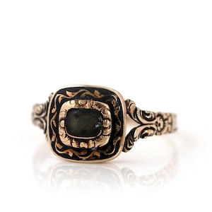 May include: A gold-toned ring with an ornate design. The ring features a rectangular, dark-colored stone set within a square frame. The frame is surrounded by black enamel and detailed gold filigree. The band has a split design with further decorative elements.