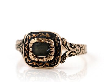 May include: A gold ring with an elaborate design. It features a rectangular, dark-coloured stone set within a square frame. The frame is surrounded by black enamel and detailed gold filigree. The band has a split design with further decorative elements.