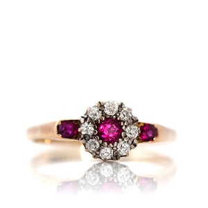 Op de afbeelding: Een gouden ring met een centrale cluster van witte diamanten rond een helder roze edelsteen. Twee kleinere roze edelstenen flankeren de centrale cluster. De ring is geplaatst tegen een witte achtergrond, wat het ontwerp benadrukt.