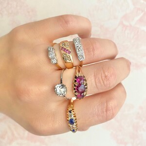 May include: A hand wearing five gold rings with various gemstones. The rings feature a pink stone, a single round diamond, a row of small diamonds, a row of small pink stones, and a row of blue stones.