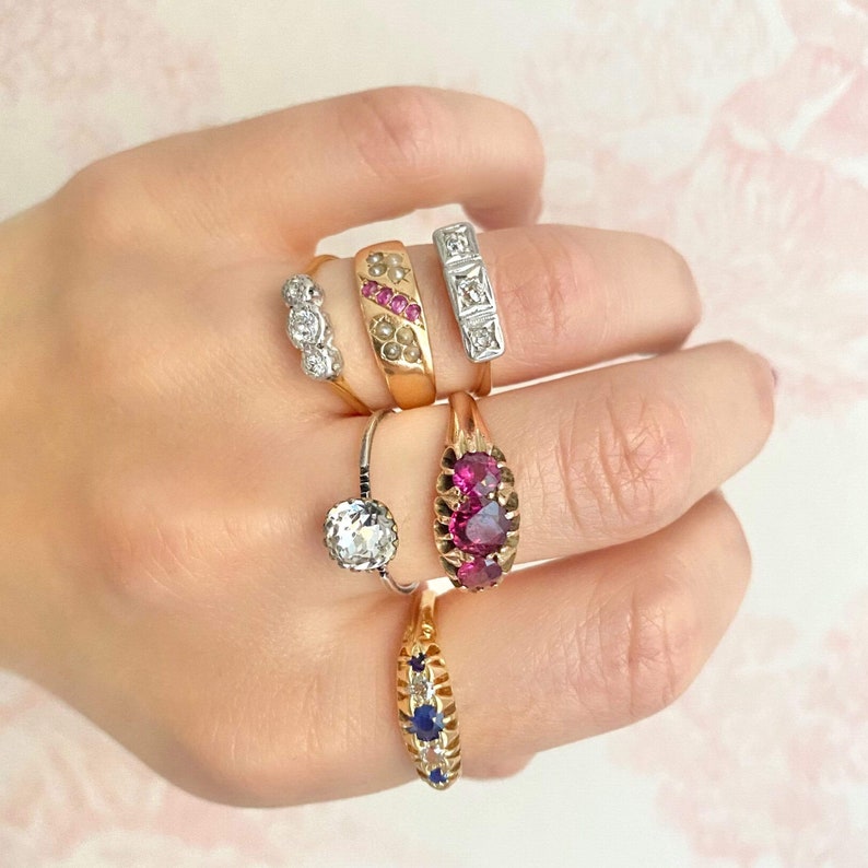 May include: A hand wearing five gold rings with various gemstones. The rings feature a pink stone, a single round diamond, a row of small diamonds, a row of small pink stones, and a row of blue stones.