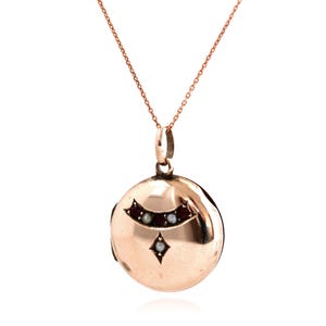Puede incluir: Collar con medallón dorado y cadena delicada. El medallón redondo presenta un diseño decorativo de media luna con pequeñas piedras preciosas rojas y blancas. Un diamante está centrado debajo.