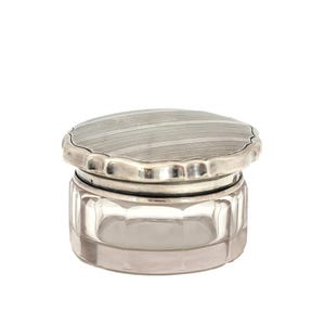 Puede incluir: Un pequeño tarro de cristal antiguo con tapa plateada. La tapa tiene un diseño a rayas y un borde festoneado. El tarro de cristal transparente tiene un diseño facetado. El tarro es probablemente para guardar cosméticos o objetos pequeños.