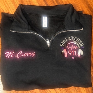 Könnte beinhalten: Schwarzes Sweatshirt mit Viertelreißverschluss und silbernem Reißverschluss. Das Sweatshirt zeigt den Namen "M. Curry" in rosa Schrift sowie ein "Dispatcher 911"-Design mit Kopfhörern und Schallwellen in Rosa und Lila.