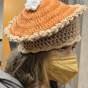 May include: A crocheted hat designed to resemble a slice of pumpkin pie. The hat features an orange top, a tan crust, and a white whipped cream accent. The hat is handmade and suitable for costume or novelty wear.