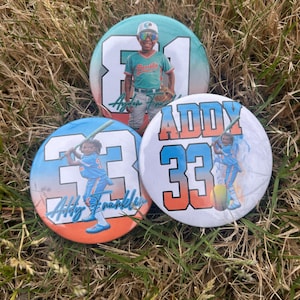 May include: Three round baseball-themed buttons with images of children in baseball uniforms. The buttons feature the numbers 81 and 33, along with the names "Aydan Franklin" and "Addy Franklin". The buttons are set on a bed of grass.
