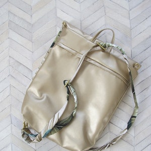 May include: A gold-coloured leather backpack with a drawstring closure and a floral patterned strap.