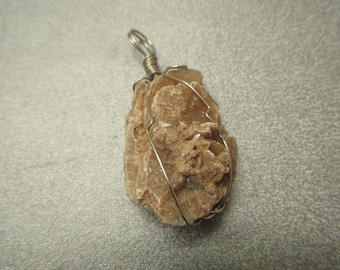 May include: A desert rose pendant, a natural formation of gypsum crystals, is wrapped in silver-colored wire. The pendant has a rough, textured surface and a light brown color, resembling a rose bloom.