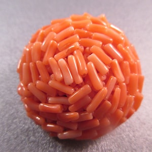 Può includere: Una sfera fatta di perline di corallo. Le perline sono piccole e cilindriche e sono disposte in un motivo casuale. Il corallo è di colore arancio chiaro.