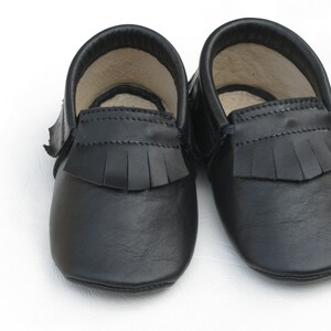 Puede incluir: Un par de mocasines de beb&eacute; de cuero negro. Los zapatos tienen una suela blanda y flecos en la parte superior. El interior de los zapatos es de color beige claro. Los zapatos est&aacute;n dise&ntilde;ados para beb&eacute;s y ni&ntilde;os peque&ntilde;os.