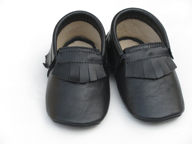 Puede incluir: Un par de mocasines de beb&eacute; de cuero negro. Los zapatos tienen una suela blanda y flecos en la parte superior. El interior de los zapatos es de color beige claro. Los zapatos est&aacute;n dise&ntilde;ados para beb&eacute;s y ni&ntilde;os peque&ntilde;os.