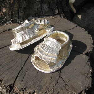 May include: A pair of gold leather sandals with fringe detailing. The sandals are sitting on a tree stump.