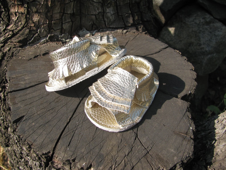 May include: A pair of gold leather sandals with fringe detailing. The sandals are sitting on a tree stump.