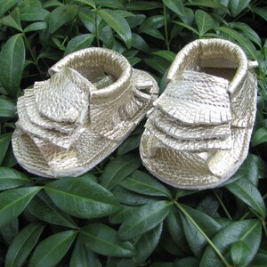 May include: A pair of gold leather baby sandals with a ruffled design. The sandals are made of soft leather and have a comfortable fit.