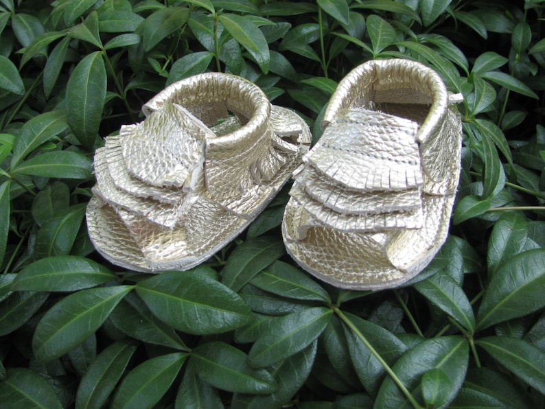 May include: A pair of gold leather baby sandals with a ruffled design. The sandals are made of soft leather and have a comfortable fit.