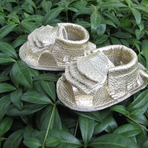 May include: A pair of gold leather baby sandals with a fringe detail. The sandals are on a green leafy background.