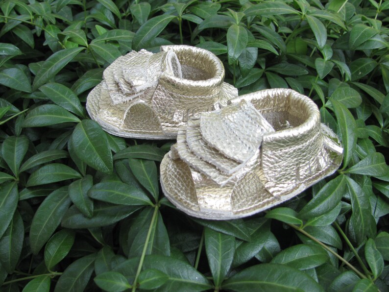 May include: A pair of gold leather baby sandals with a fringe detail. The sandals are on a green leafy background.