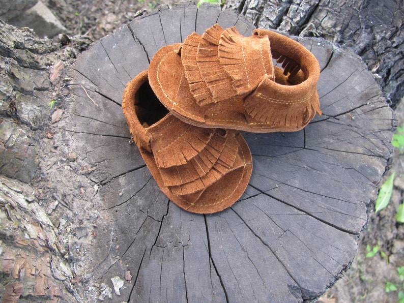 May include: A pair of brown suede baby moccasins with fringe detailing. The moccasins are sitting on a tree stump.