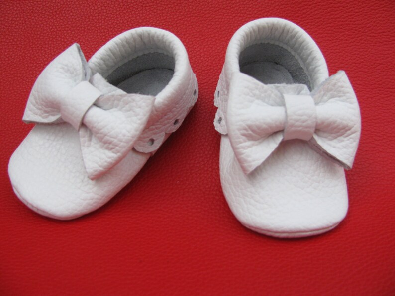 May include: A pair of white leather baby moccasins with a bow on the front.