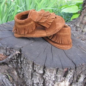 May include: A pair of brown suede baby moccasins with fringe detailing. The moccasins are sitting on a tree stump.