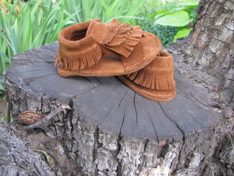 May include: A pair of brown suede baby moccasins with fringe detailing. The moccasins are sitting on a tree stump.