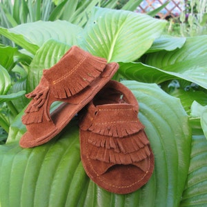 May include: Two pairs of brown suede sandals with fringe detailing. The sandals are on a green leafy background.