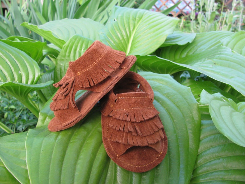 May include: Two pairs of brown suede sandals with fringe detailing. The sandals are on a green leafy background.