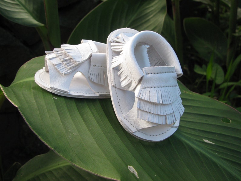 May include: A pair of white leather sandals with fringe detailing. The sandals are on a green leaf.