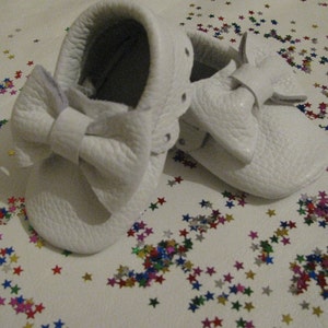 May include: A pair of white leather baby moccasins with a large white bow on the front. The moccasins are on a white surface with colorful confetti.