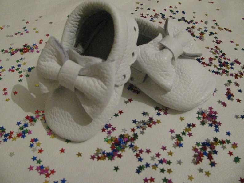 May include: A pair of white leather baby moccasins with a large white bow on the front. The moccasins are on a white surface with colorful confetti.