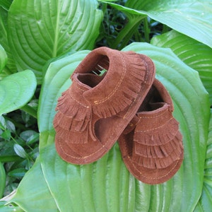 May include: A pair of brown suede baby moccasins with fringe detailing. The moccasins are on a green leafy background.