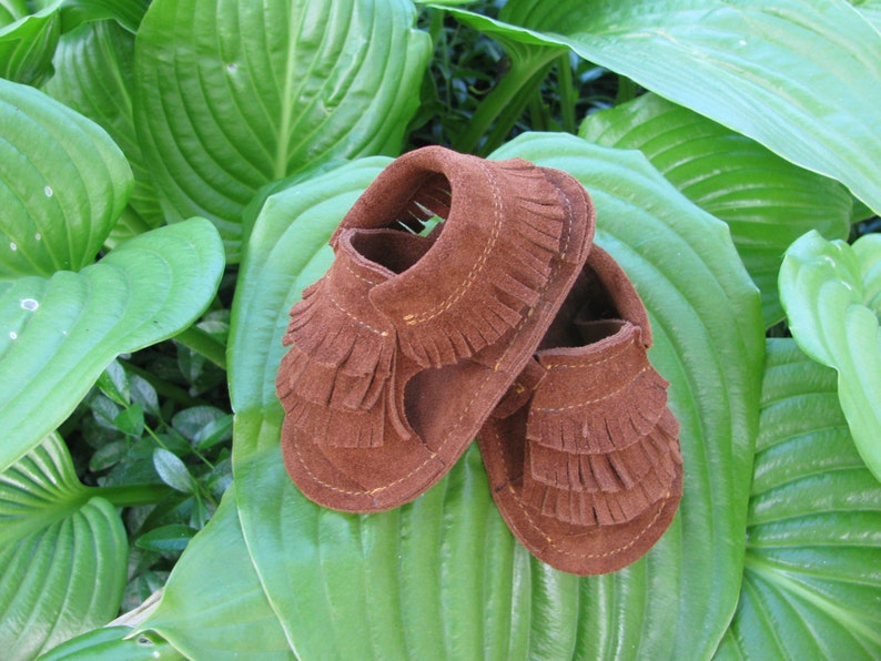 May include: A pair of brown suede baby moccasins with fringe detailing. The moccasins are on a green leafy background.