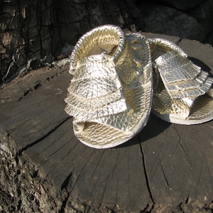May include: A pair of gold sandals with a ruffled design. The sandals are made of a textured material and have a strap around the ankle.